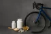 Compléments alimentaires pour cyclistes posés près de la roue avant d’un vélo de route bleu, sur un fond gris.